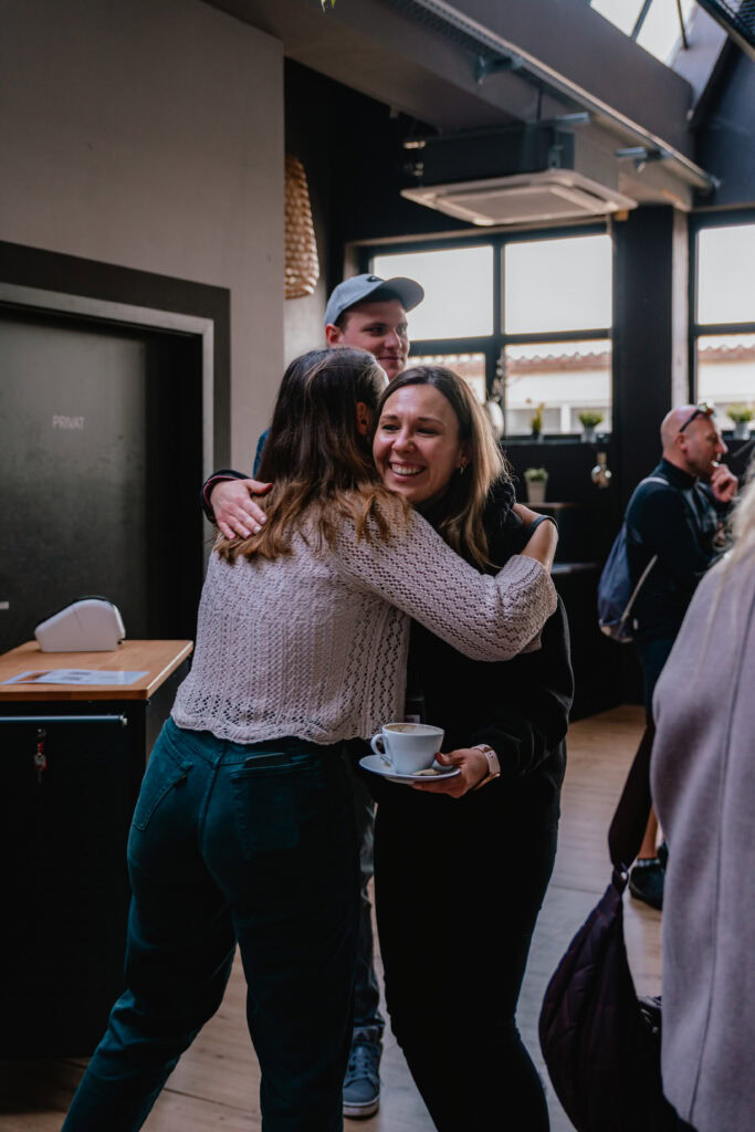 Zwei Frauen umarmen sich, eine hält eine Tasse Kaffee, im Hintergrund weitere Personen.
