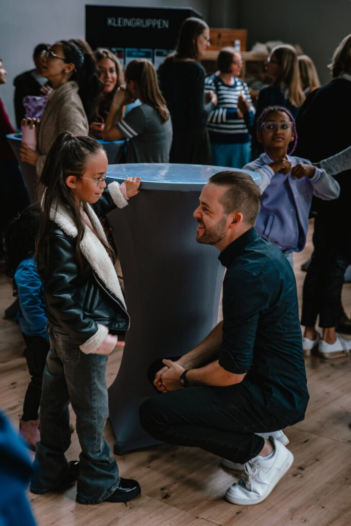 Ein Mann kniet vor einem Mädchen, beide sprechen sich an. Im Hintergrund stehen weitere Kinder und E.