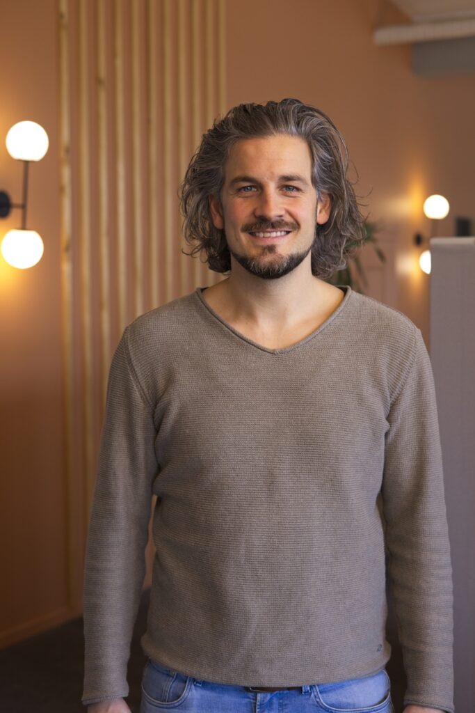 Freundlicher Mann mit lockigem Haar und Bart in einem modernen, hell erleuchteten Innenraum, der für Veranstaltungen oder Treffen bei ICF Basel geeignet ist.