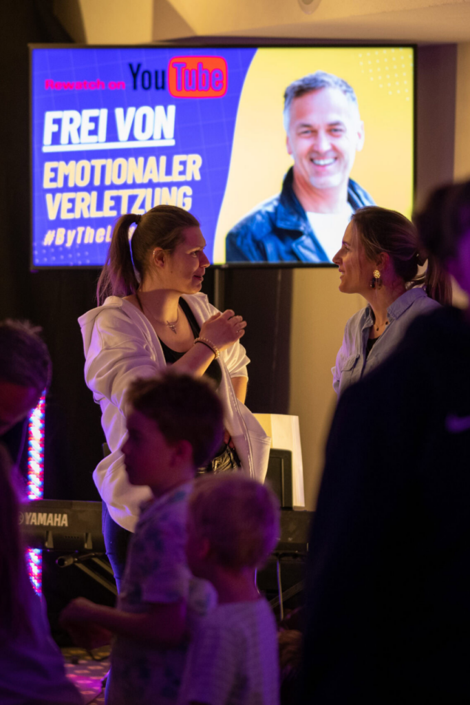 Zwei Frauen sprechen sich bei einer Veranstaltung vor einem Bildschirm mit einem Mann.