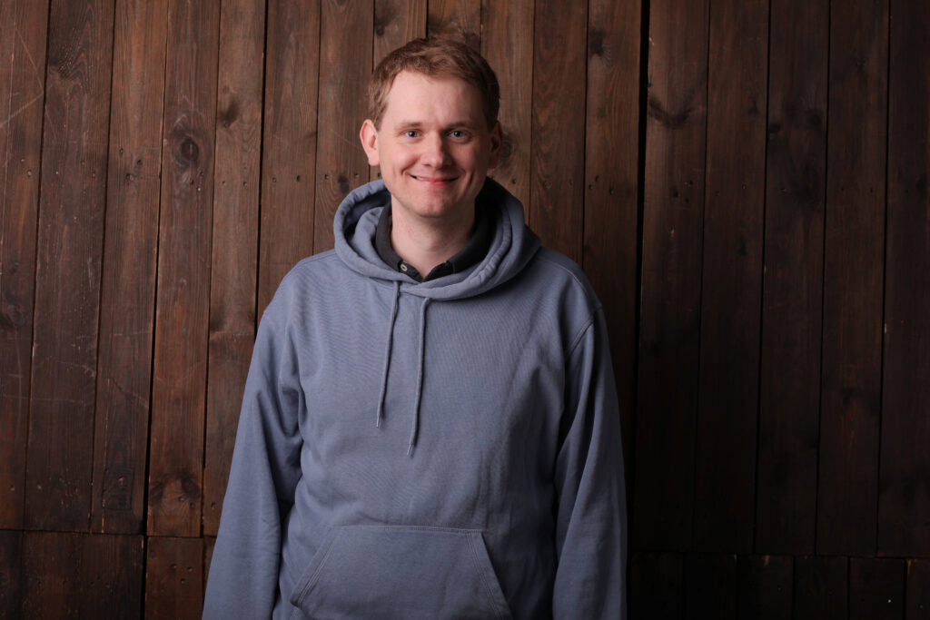 Freundlicher junger Mann in grauem Hoodie vor Holzpaneel, lächelnd, für christliche Gemeinschaft Rhein-Main, geeignet für Kirche, Glaube, Spiritualität, Gemeinschaft.
