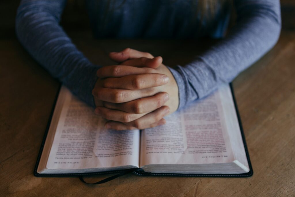Glaubende Hände ruhen auf einer aufgeschlagenen Bibel, symbolisieren Glauben, Gebet und spirituelle Verbundenheit im christlichen Glauben.