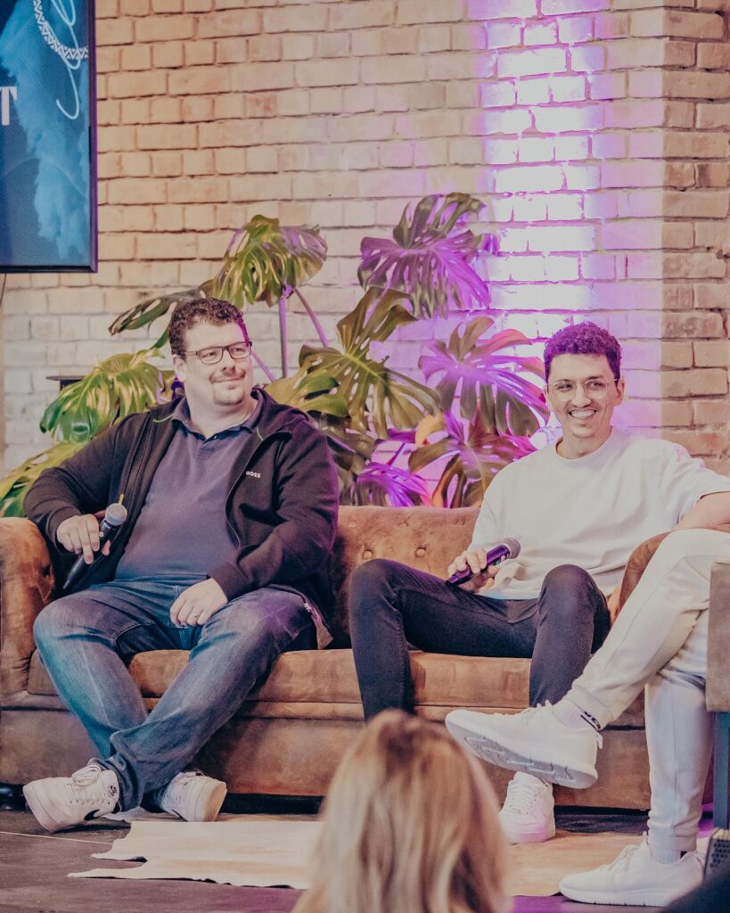 Alt: Zwei junge Männer beim Gemeindetreff in Mannheim sitzen auf einem Sofa, lachen und sprechen in einem modernen, urbanen Raum mit Pflanzen und Backsteinmauer.