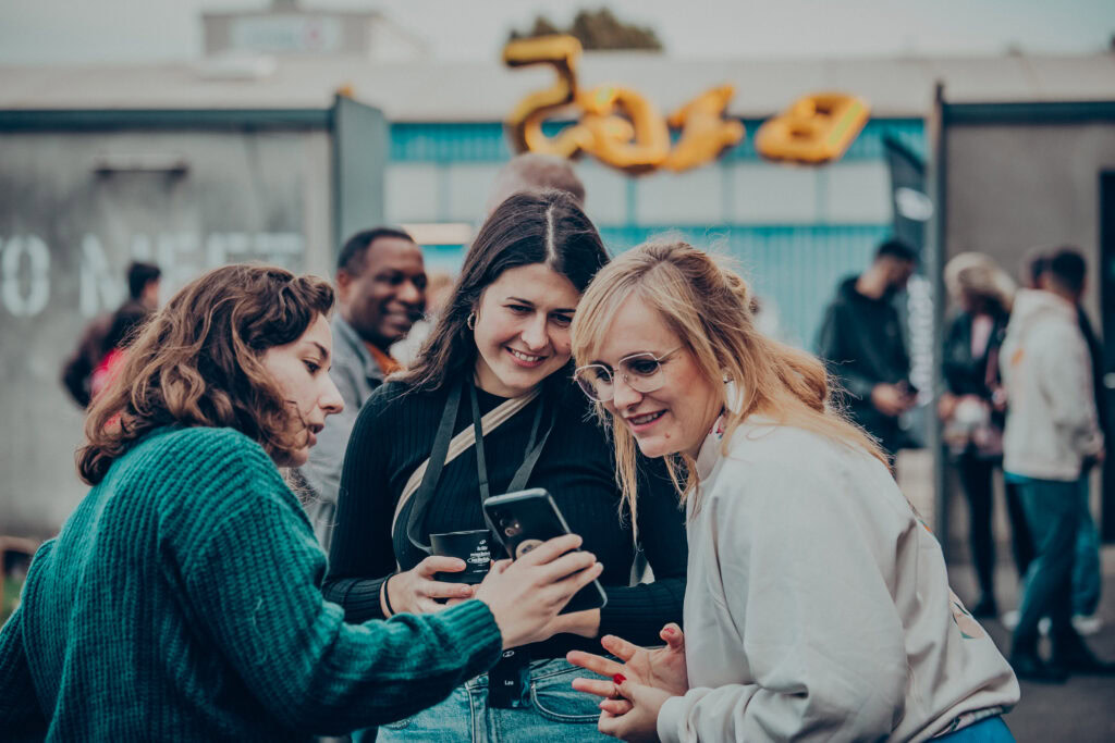 Fröhliche Frauen beim Austausch, schauen auf Smartphone, tragen festliche Kleidung, genießen Gemeinschaft bei der Veranstaltung in Mannheim, Deutschland.