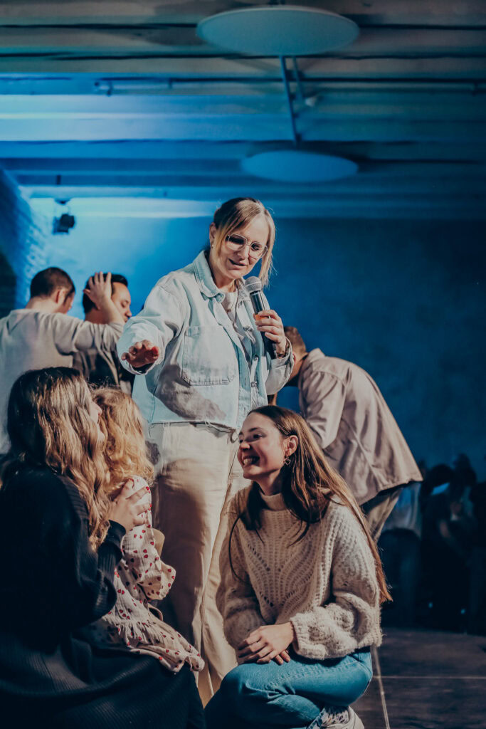 Alt: Begeisterte Gemeindemitglieder bei einem Gemeinschaftsabend in der ICF Kirche Mannheim, engagierte Predigerin spricht mit jungen Teilnehmern, moderne Atmosphäre, Gemeinschaft und Glauben im Mittelpunkt.