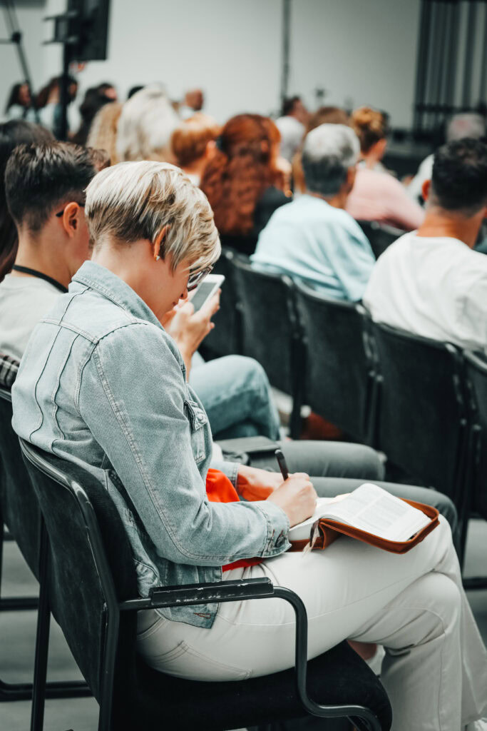 Alt-Text: Jugendliche und Erwachsene sitzen während einer Predigt im Gottesdienst in Mannheim, einige notieren, andere schauen auf ihr Smartphone.